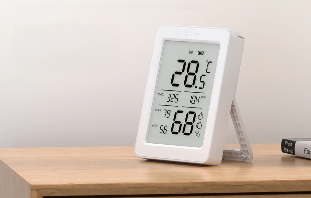 Senzorul Sonoff AirGuard TH este așezat pe un raft de lemn și afișează temperatura și umiditatea pe un ecran LCD mare.