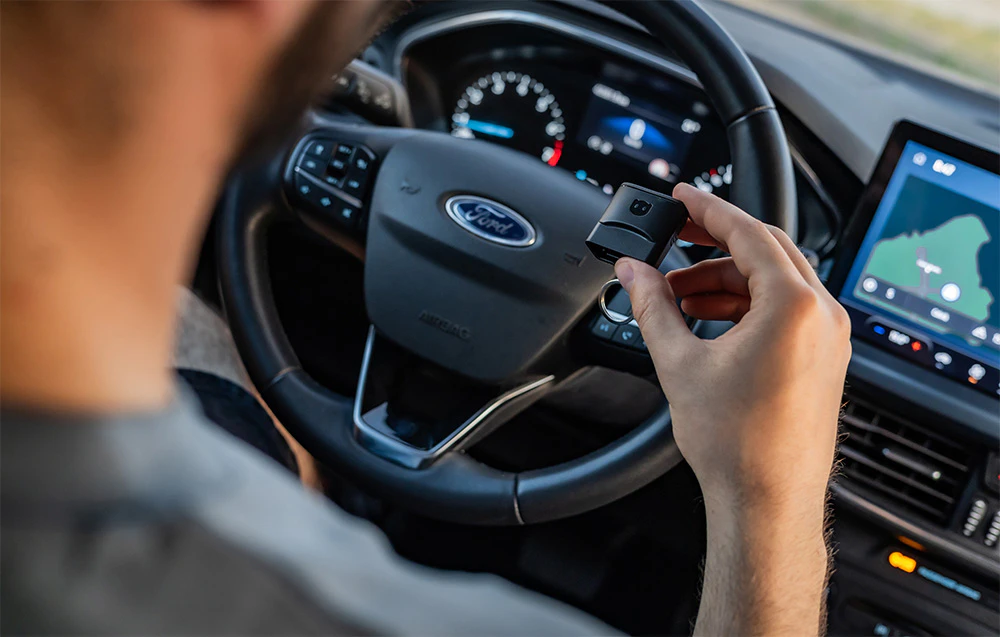 Șofer ținând în mână adaptorul Carly OBD2 în interiorul unei mașini Ford, cu ecranul media vizibil
