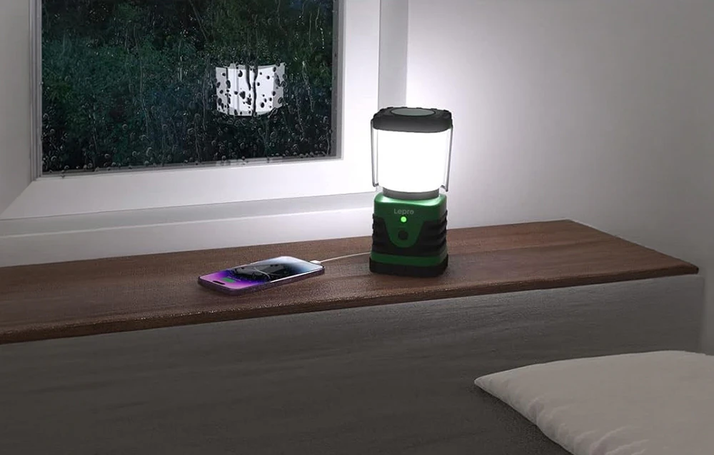O lampă de camping stă pe o masă de lemn, încărcând un smartphone conectat printr-un cablu; în fundal, se vede o fereastră cu picături de ploaie.