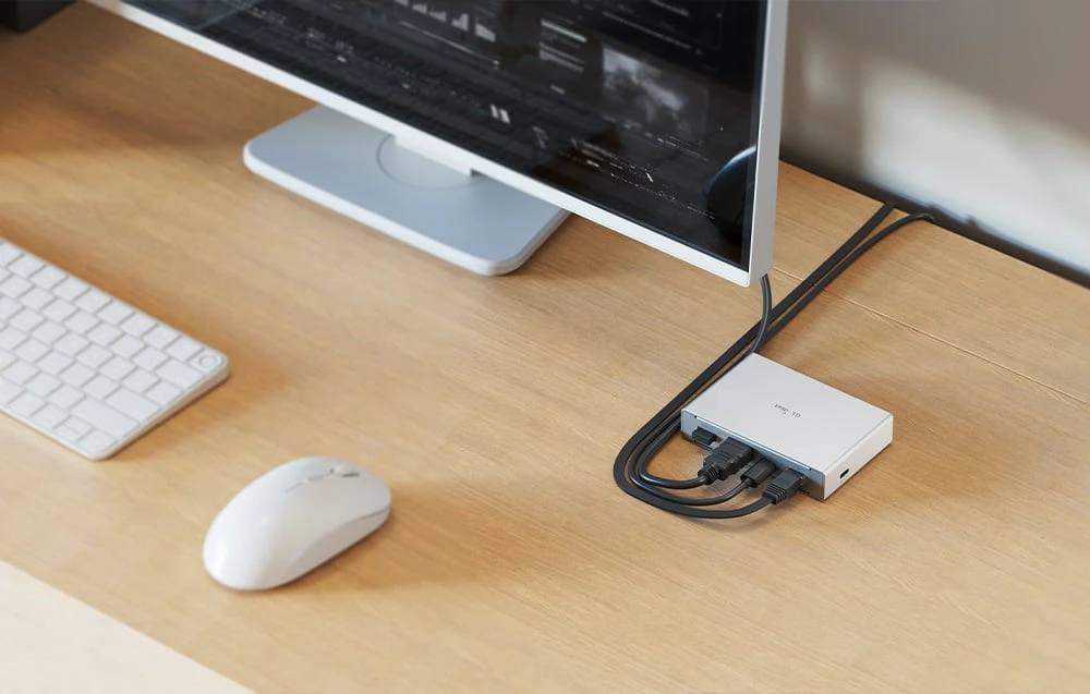 Un comutator KVM argintiu conectat prin cabluri pe un birou din lemn; lângă acesta se văd un mouse și o tastatură albe.