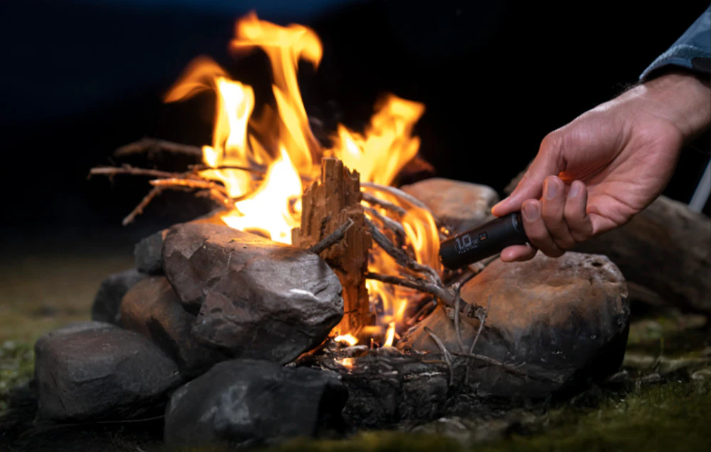 O mână ține o pompă mică, neagră, îndreptată spre un foc de tabără aprins, făcut din pietre și crengi