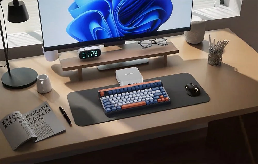 Un birou cu un mini PC, monitor, tastatură, mouse, carte și alte obiecte