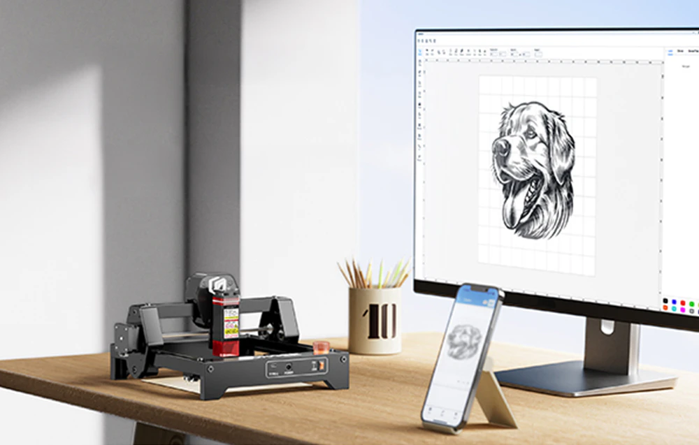Gravor laser pe un birou conectat la un computer, cu un desen cu un câine vizibil pe monitor și smartphone