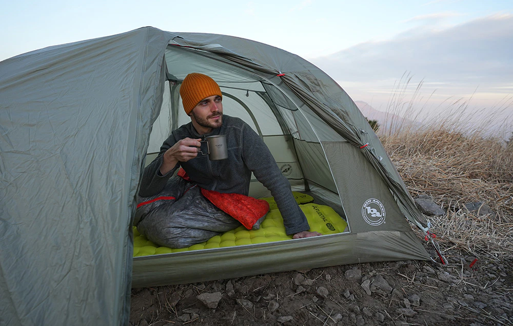 Un drumeț stă într-un cort pe un covoraș de camping desfăcut, bând cafea dintr-o cană de oțel în timp ce privește munții