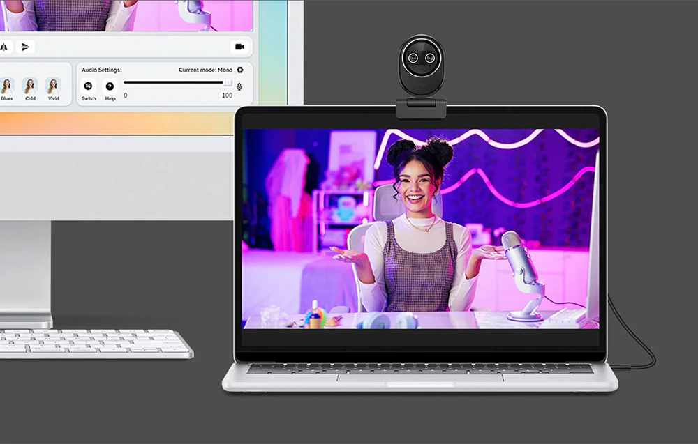 Un laptop cu o cameră web atașată; ecranul arată o femeie care prezintă o transmisiune live, cu un monitor și o tastatură lângă ea