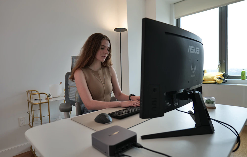 O femeie cu părul lung stă la un birou și tastează la o tastatură, având în fața ei un mini PC și un monitor