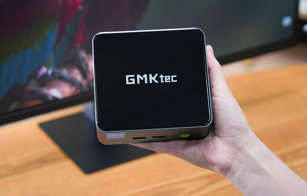 Computerul compact GMKtec M3 Pro ținut într-o mână pe fundalul unui monitor