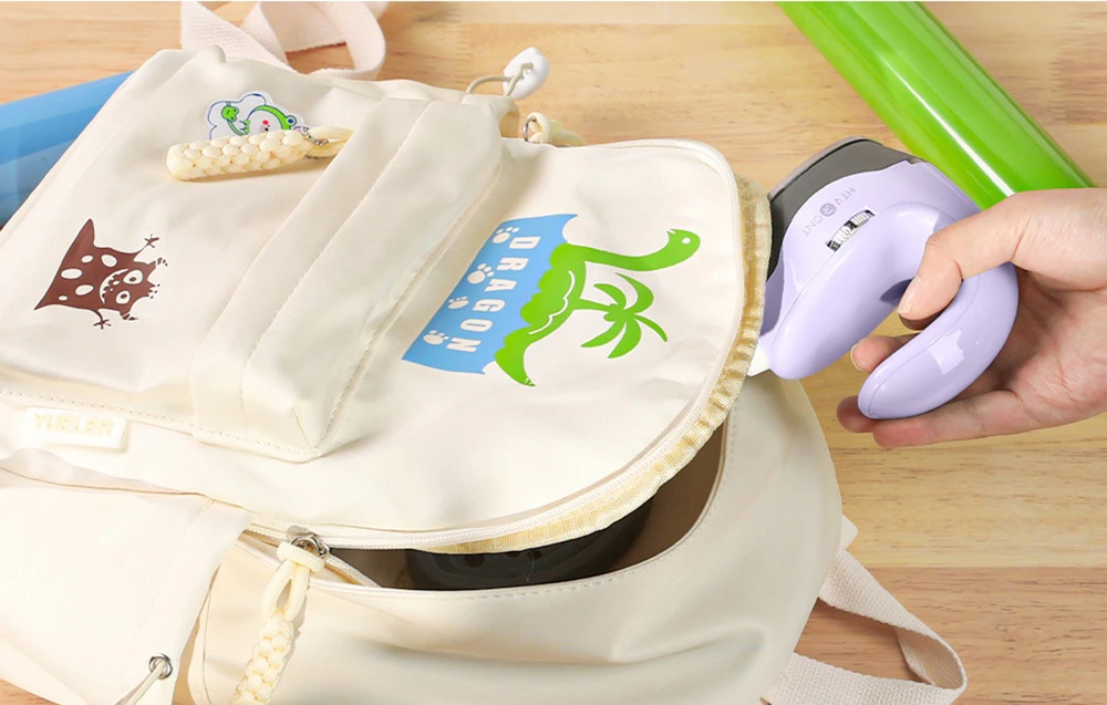 O mână introduce presa de căldură violetă într-un rucsac alb deschis, cu un desen cu un dinozaur verde.