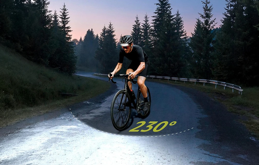 Un ciclist care poartă cască și are lanterna frontală aprinsă pedalează pe un drum asfaltat. Cuvintele „230 de grade” în galben sunt vizibile pe drum.