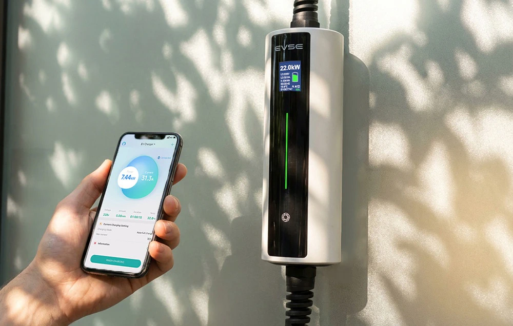 Un smartphone ținut în mână cu aplicația deschisă, un încărcător pentru vehicule electrice pe perete lângă acesta
