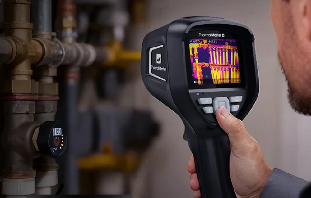 O mână ține o cameră termică Thermal Master, al cărei ecran afișează o imagine termică a unui sistem de țevi