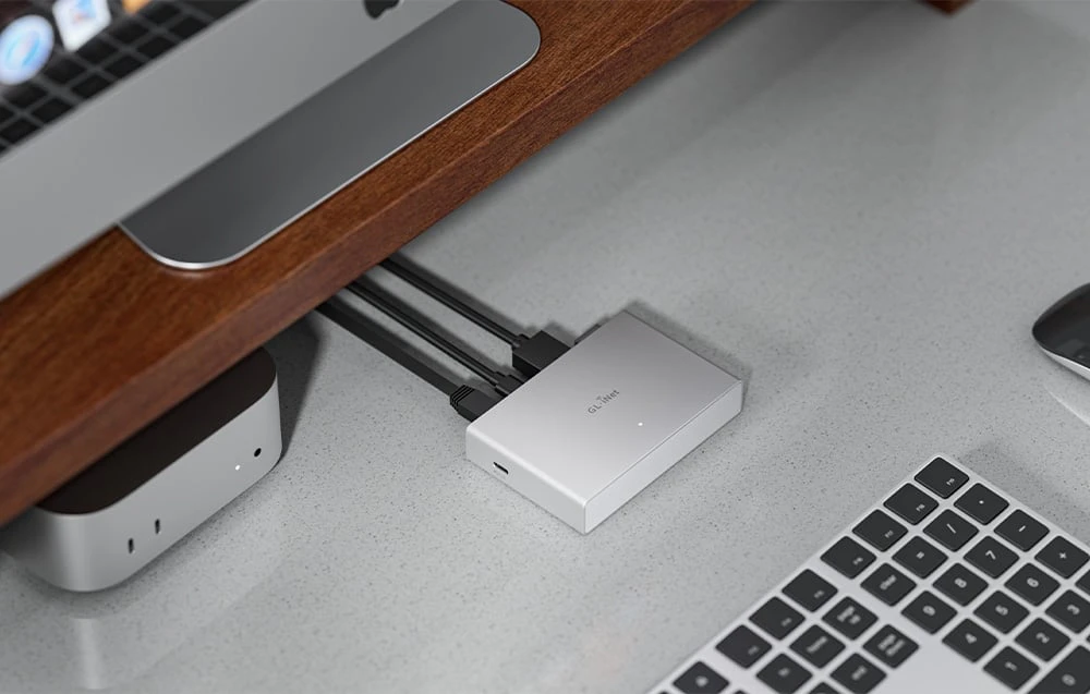 Un comutator KVM argintiu pe un blat de culoare deschisă, așezat sub un raft din lemn lângă un Mac Mini.