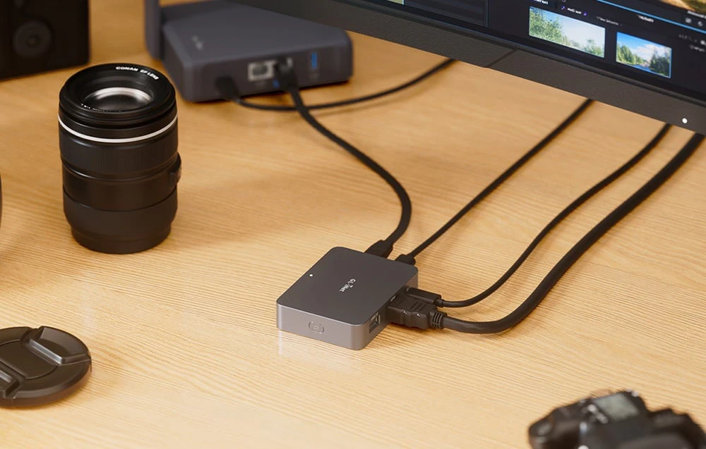 Un comutator KVM gri așezat pe un birou din lemn, conectat prin mai multe cabluri negre sub un monitor de computer