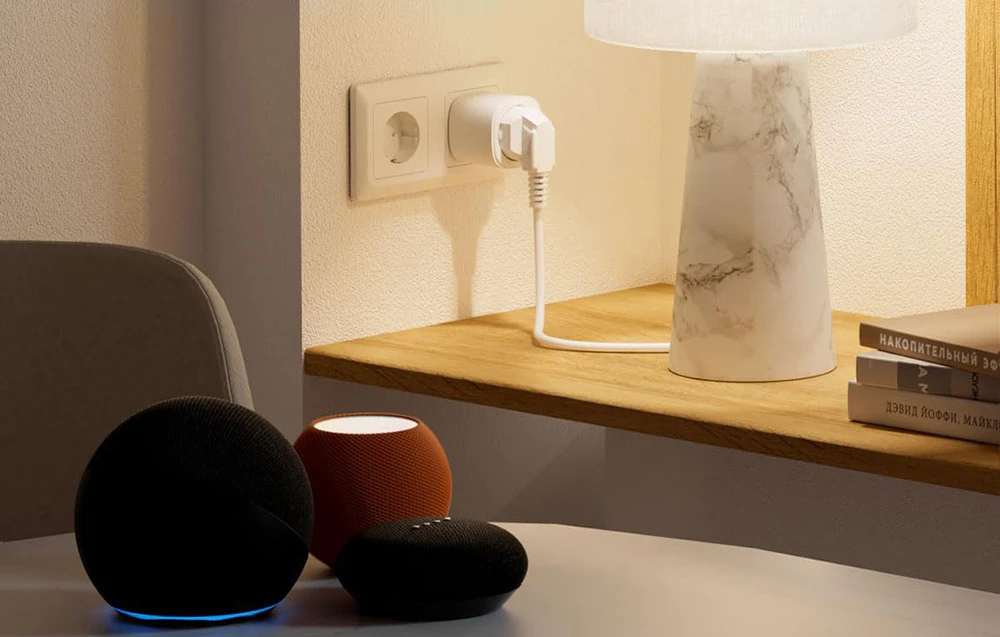 Trei difuzoare inteligente Amazon Echo în culorile negru, portocaliu și negru, așezate pe o masă în fața unei prize inteligente conectate la o priză de curent și a unei lămpi pe un raft din lemn