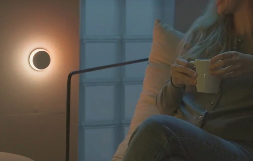 Lampă de noapte Yeelight Vega care luminează cu lumină caldă pe perete, lângă o persoană așezată care ține o cană.