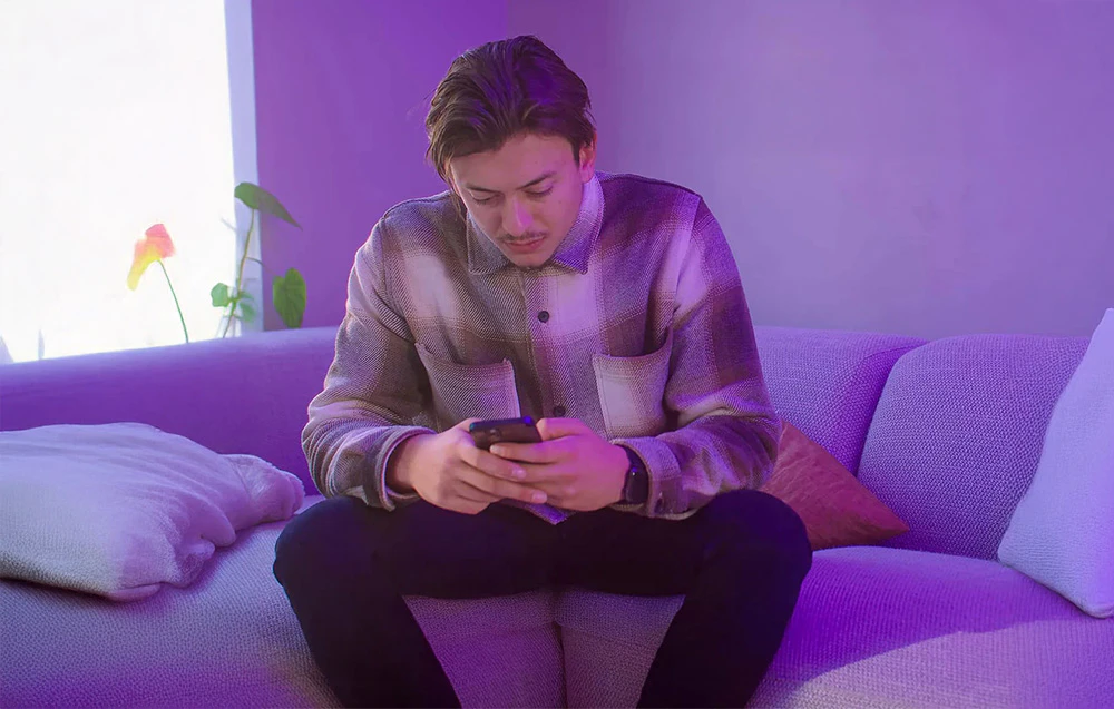 Un tânăr stă pe o canapea mov și controlează încuietoarea prin intermediul unei aplicații mobile pe un smartphone