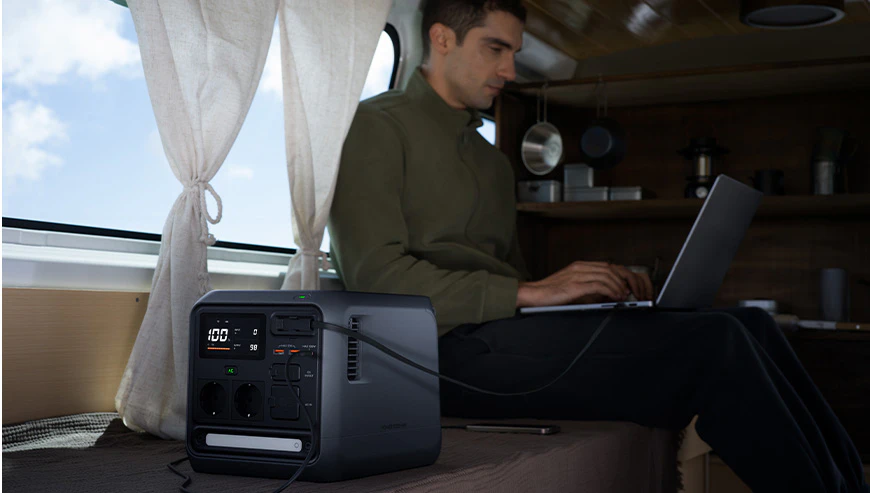 Un bărbat stă în interiorul unei rulote și lucrează la un laptop, care este conectat printr-un cablu la stația de alimentare DJI Power 1000 Mini așezată lângă el pe pat