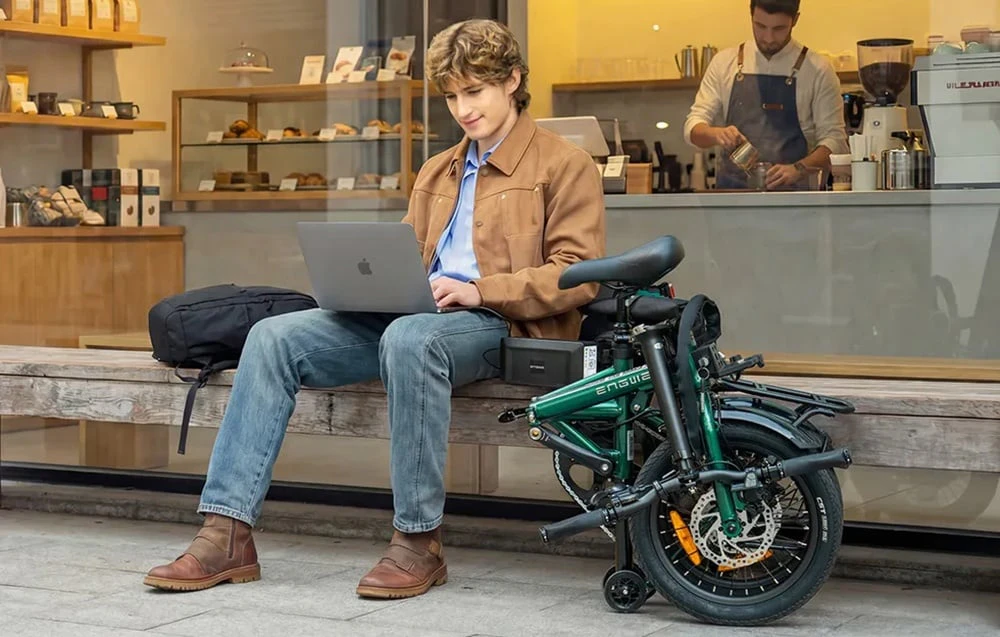 Un bărbat lucrează la un laptop în fața unei cafenele, cu o bicicletă electrică pliabilă lângă el