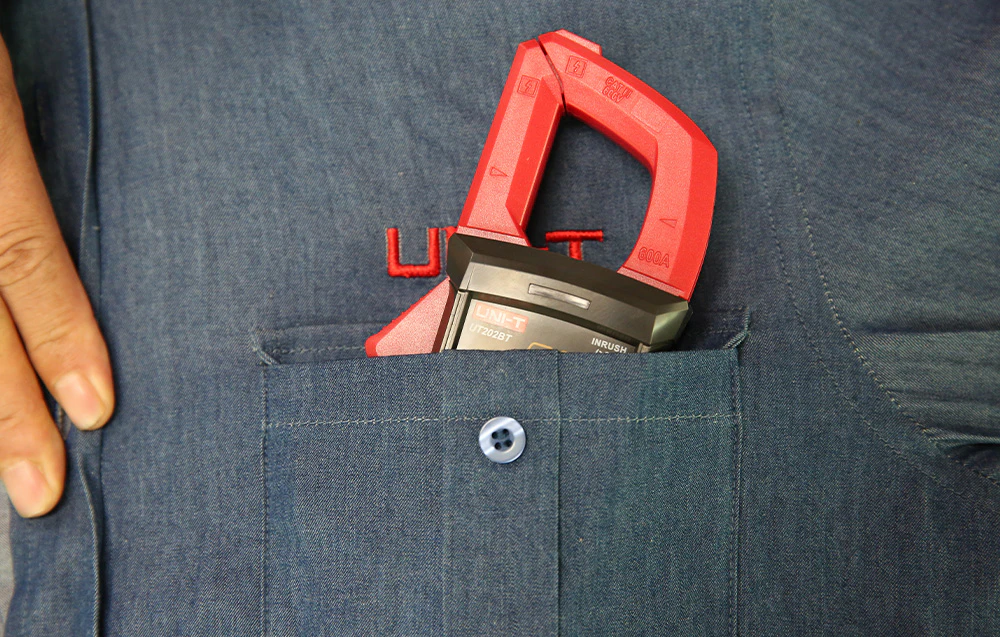 Un clește ampermetric UNI-T roșu în buzunarul unei cămăși din denim, lângă o mână.