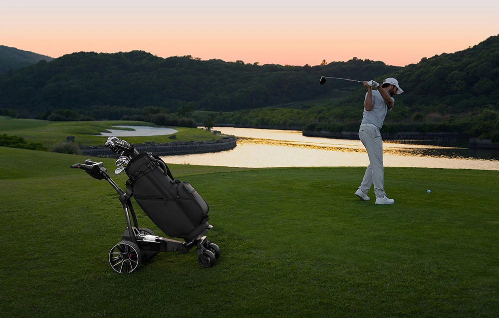 Un jucător de golf lovește mingea la apusul soarelui, cu un cărucior de golf automat cu un sac de golf lângă el