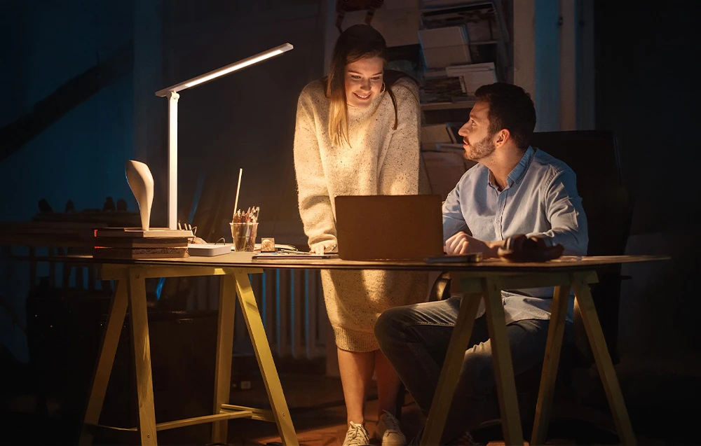 O femeie și un bărbat stau lângă un birou într-o cameră întunecată, uitându-se la ecranul unui laptop iluminat de o lampă de birou albă.