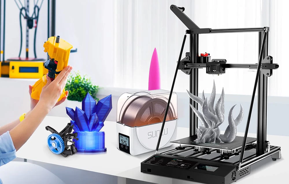 Imprimantă 3D în funcțiune lângă uscătorul SUNLU și modele 3D