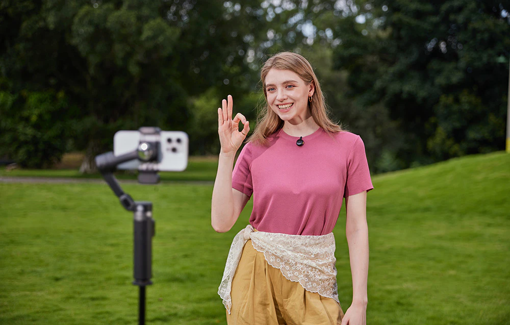 O femeie zâmbește la cameră și face un gest de „OK” în timp ce înregistrează cu un smartphone montat pe un gimbal.
