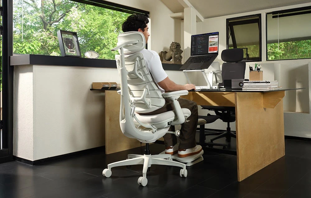 O persoană stă așezată într-un scaun ergonomic alb la un birou, folosind un laptop așezat pe un suport