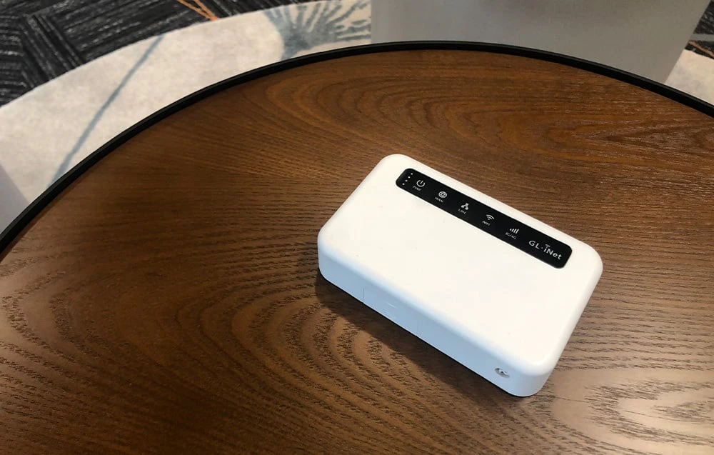 Un router alb de dimensiuni mici așezat pe o măsuță de cafea din lemn de culoare închisă; se vede panoul cu pictogramele și logo-ul GL.iNet.