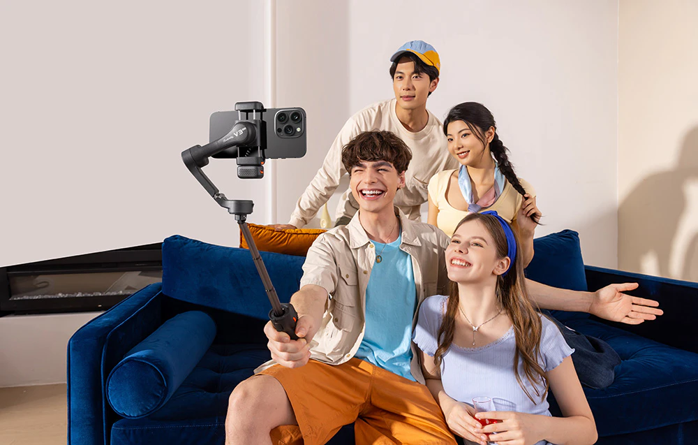 Patru tineri își fac un selfie cu un smartphone montat pe un gimbal într-o cameră de zi.