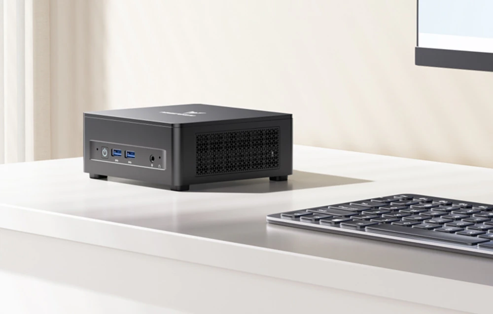 Un mini PC negru pe un birou alb, lângă o tastatură, cu porturi USB și audio vizibile.