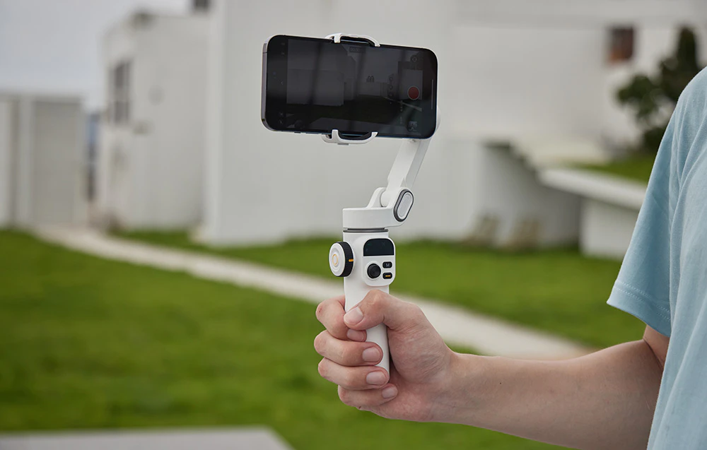 O mână ținând un gimbal alb cu un smartphone în poziție orizontală în timp ce înregistrează în aer liber.