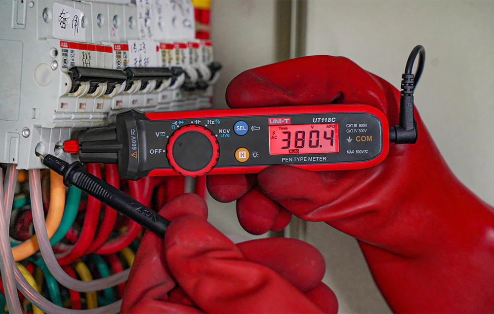 Măsurarea tensiunii într-un tablou electric folosind un multimetru UNI-T ținut de un operator care poartă mănuși de protecție roșii.