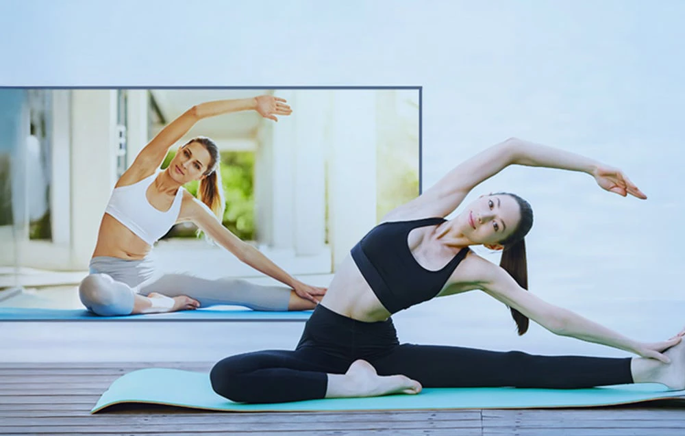 O femeie practică yoga pe o saltea, imitând mișcările instructorului vizibil într-o imagine mare proiectată pe perete