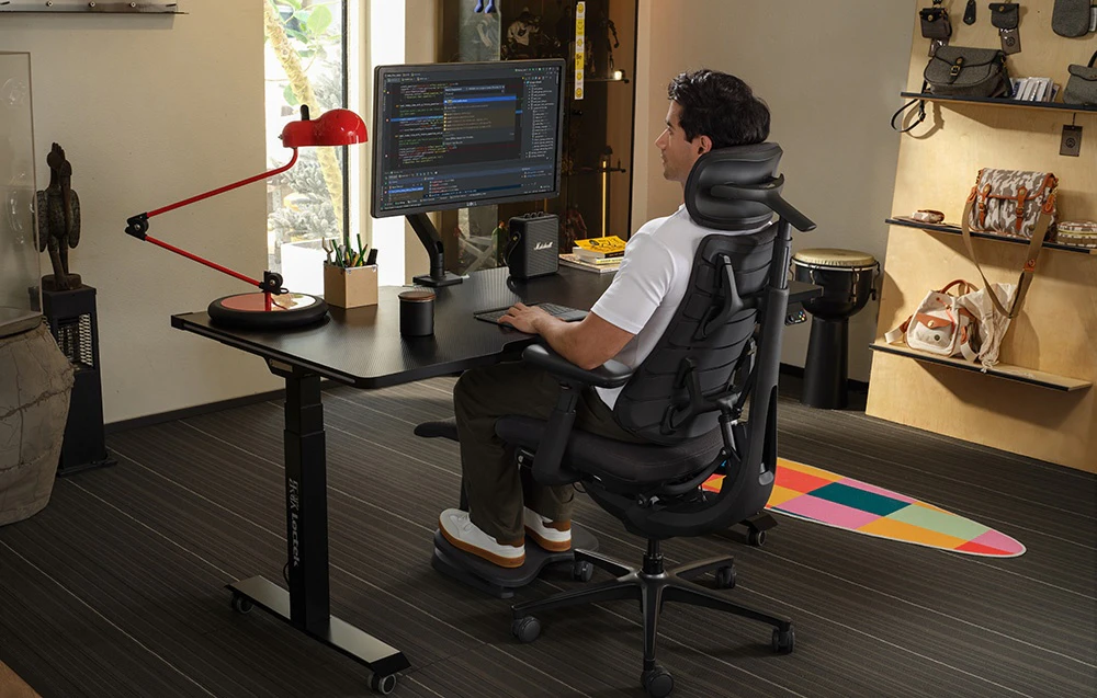 Un bărbat stă așezat într-un scaun ergonomic la un birou, lucrând la un computer de birou