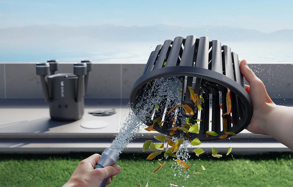 Unele mâini țin coșul de filtrare al robotului, clătindu-l cu un jet de apă provenit dintr-un furtun de grădină aflat la marginea piscinei