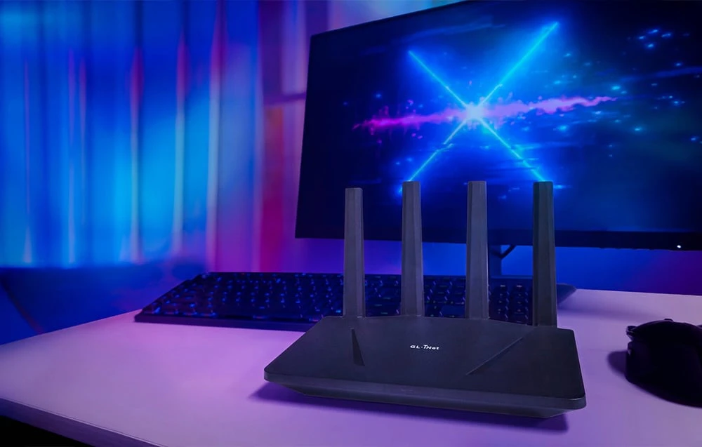 Router negru așezat pe un birou, având ca fundal un monitor pornit care afișează elemente grafice albastre