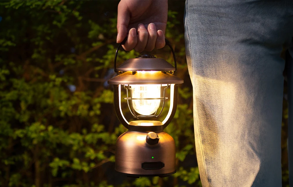 O persoană ține în mână o lampă de camping din cupru, pe fundalul unor tufișuri verzi