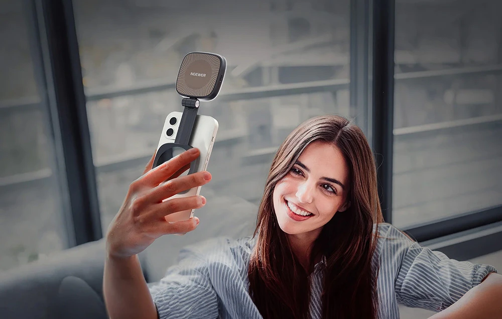O femeie zâmbitoare care își face un selfie cu smartphone-ul, folosind o lampă LED Neewer magnetică, extensibilă, montată pe un braț reglabil.