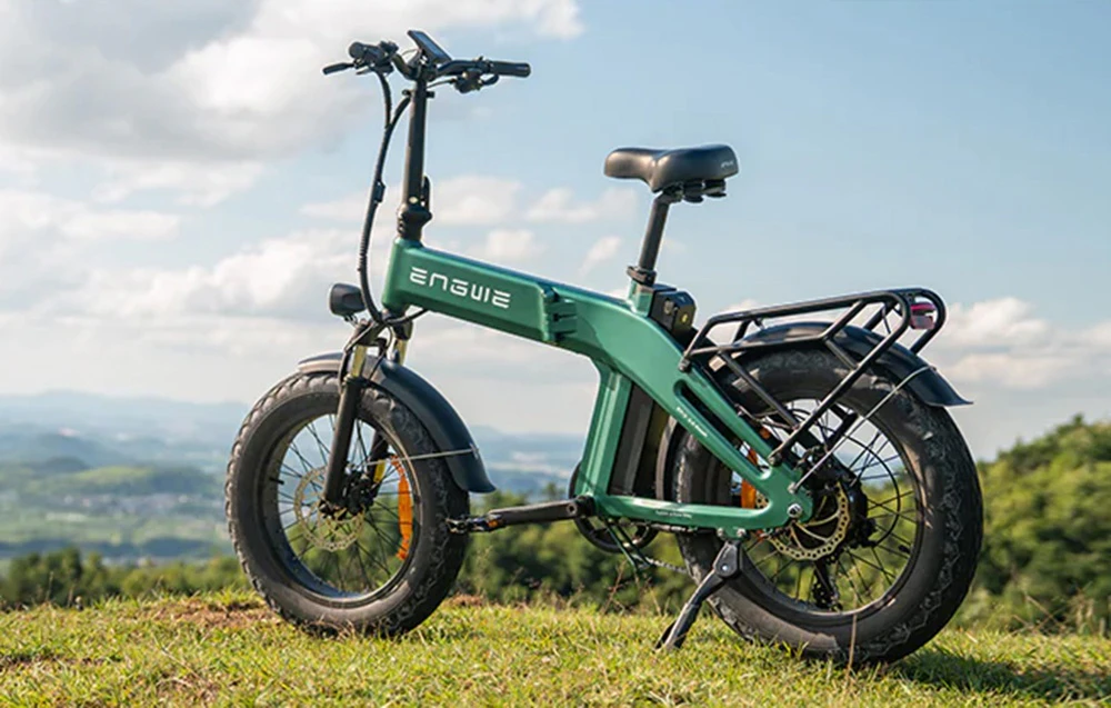 O bicicletă electrică pliabilă verde stă pe iarbă, având în fundal un peisaj montan