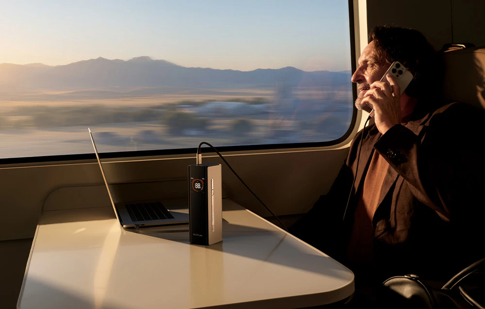 Un bărbat stă într-un tren și vorbește la telefon. Lângă el, pe masă, se află un laptop conectat la un display powerbank.