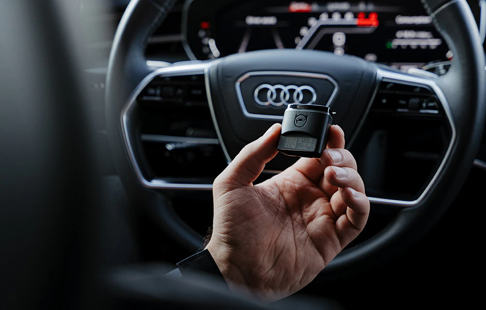 Adaptor de diagnosticare Carly OBD2 ținut în fața volanului cu sigla Audi în cockpit digital