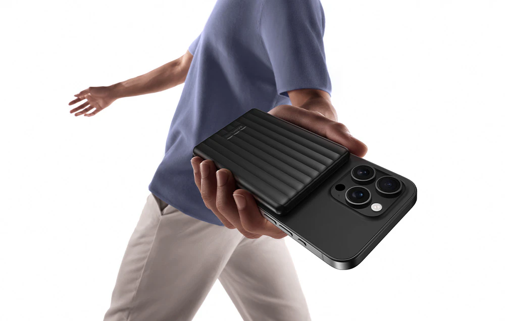 Un bărbat în cămașă mov și pantaloni de culoare deschisă ține un smartphone conectat la un powerbank magnetic.