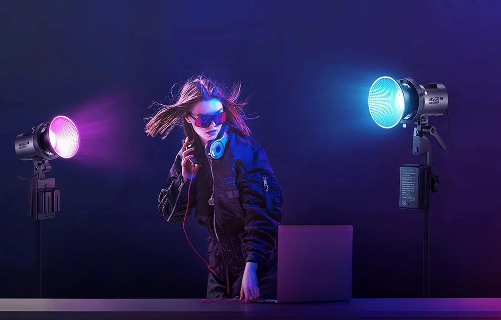 O femeie cu ochelari de soare stă lângă un laptop, iluminată din ambele părți de lumina colorată a lămpilor.
