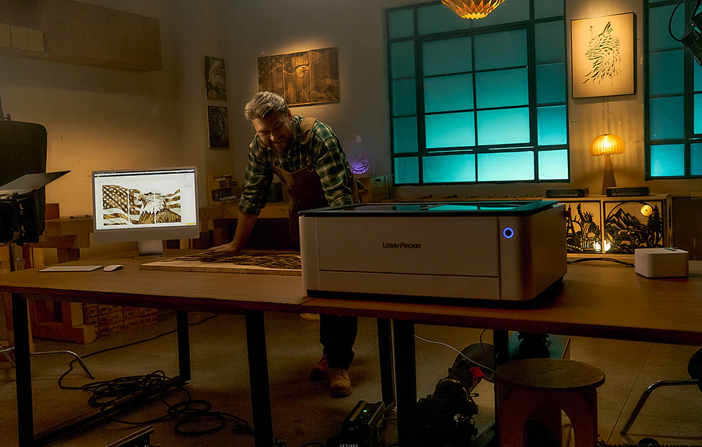 Un bărbat în șorț se uită la o placă de lemn lângă mașina LaserPecker într-un atelier slab iluminat