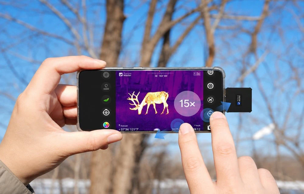 Smartphone-ul din mâini afișează o imagine termică a unui cerb cu zoom de 15x. În fundal se vede o pădure iarna.