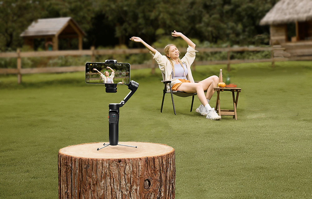 O femeie stând pe un scaun pe iarbă, înregistrată de un gimbal fixat pe un trunchi de copac.
