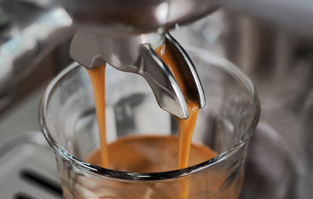 Prim-plan al duzei mașinii de espresso, din care un jet de cafea fierbinte cu spumă groasă (crema) curge într-un pahar.