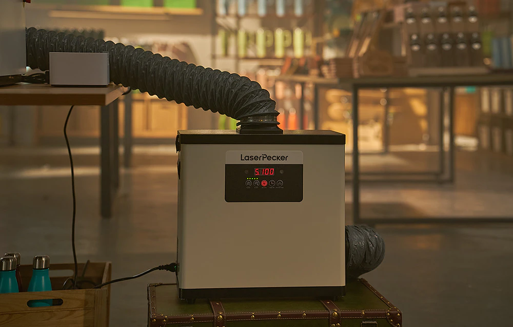 Dispozitiv LaserPecker cu o țeavă de ventilație conectată plasată pe o cutie într-un magazin sau atelier