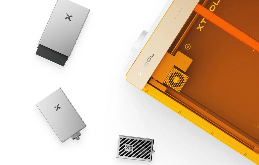 Vedere de sus a plotterului xTool cu capacul său portocaliu, alături de modulele laser interschimbabile și accesoriile de gravare așezate lângă acesta